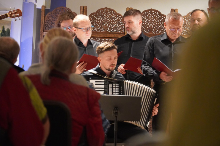 Mężczyzna gra na harmonii; za nim siedzą członkowie chóru w czarnych strojach, trzymający czerwone teczki z nutami.