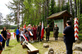 Grupa młodzieży z biało-czerwonymi flagami stoi na skarju lasu i słucha dorosłej kobiety.