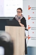 Prelegentka konferencji naukowej przemawia do mikrofonu przy mównicy z laptopem. W tle widoczne są banery informacyjne z tytułem konferencji „Opozycyjne formy zaangażowania kobiet w systemach autorytarnych i totalitarnych XX wieku” oraz logotypy Instytutu Pileckiego i programu Ministerstwa Nauki „Wektory Nauki”.
