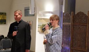 Kobieta przemawia do zgromadzonych z mikrofonem; obok niego stoi duchowny z mikrofonem.