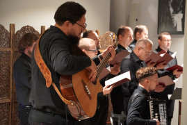 Mężczyzna gra na gitarze; za nim siedzą członkowie chóru w czarnych strojach, trzymający czerwone teczki z nutami.