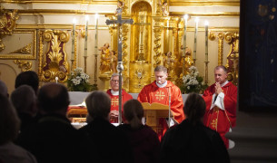 Msza w ozdobionym złoceniami kościele; trzech duchownych w czerwonych szatach liturgicznych sprawuje nabożeństwo przed ołtarzem, a w tle modlą się wierni.