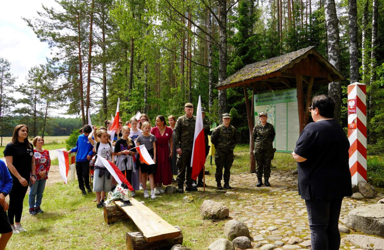 Grupa młodzieży z biało-czerwonymi flagami stoi na skarju lasu i słucha dorosłej kobiety. 