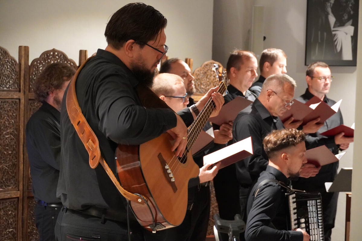 Mężczyzna gra na gitarze; za nim siedzą członkowie chóru w czarnych strojach, trzymający czerwone teczki z nutami.
